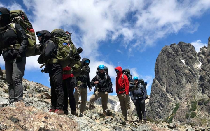 A group of people wearing backpacks make their way over a rocky landscape. There are tall rock formations in the background.